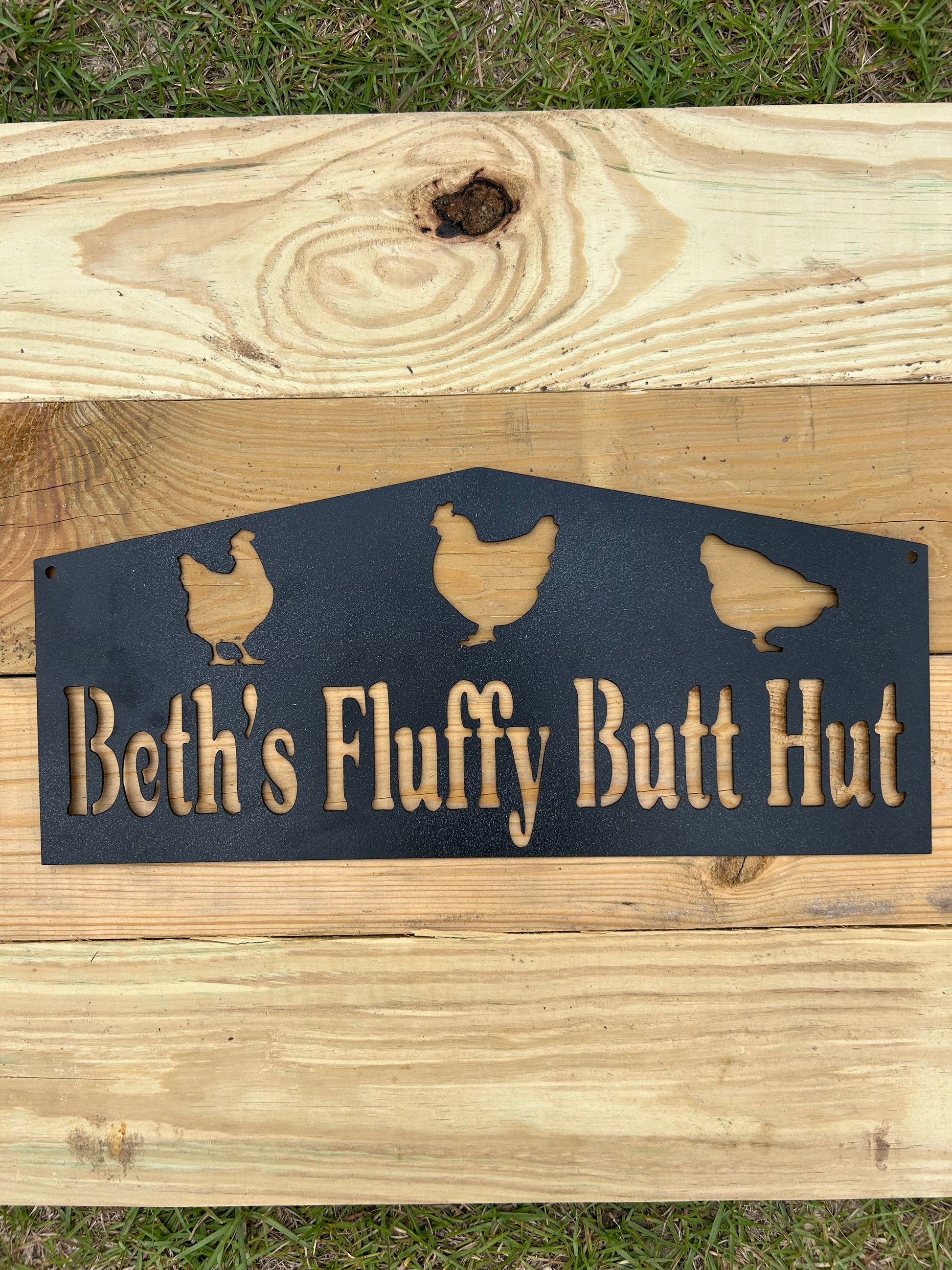 metal sign with 'Beth's Fluffy Butt Hut' text and hen illustrations on a wooden background.